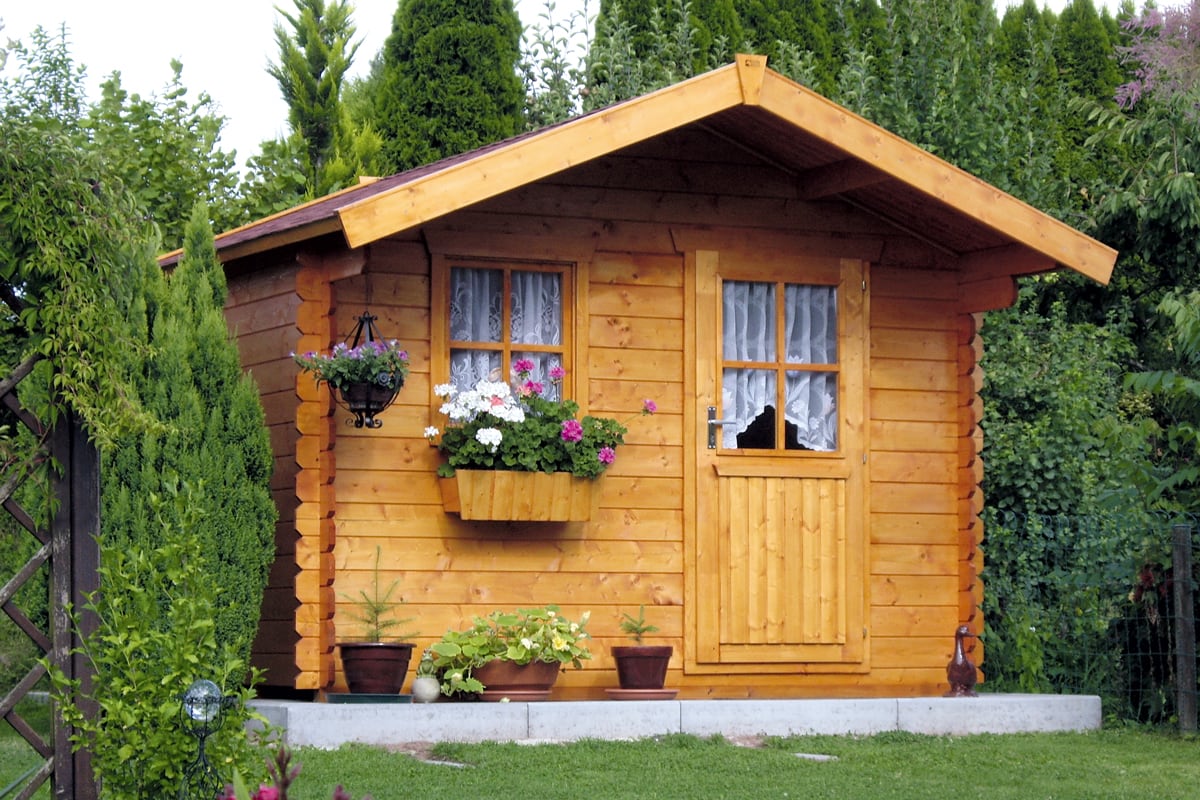Kleines Gartenhaus aus Holz mit zwei Fenstern und einer Tür, umgeben von grünen Pflanzen und Rasen