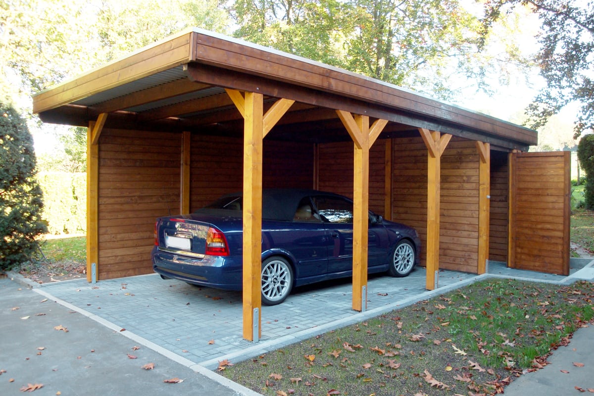 Offener Carport aus hellem Holz mit Flachdach, seitlicher Abstellkammer, darin ein blauer Pkw.