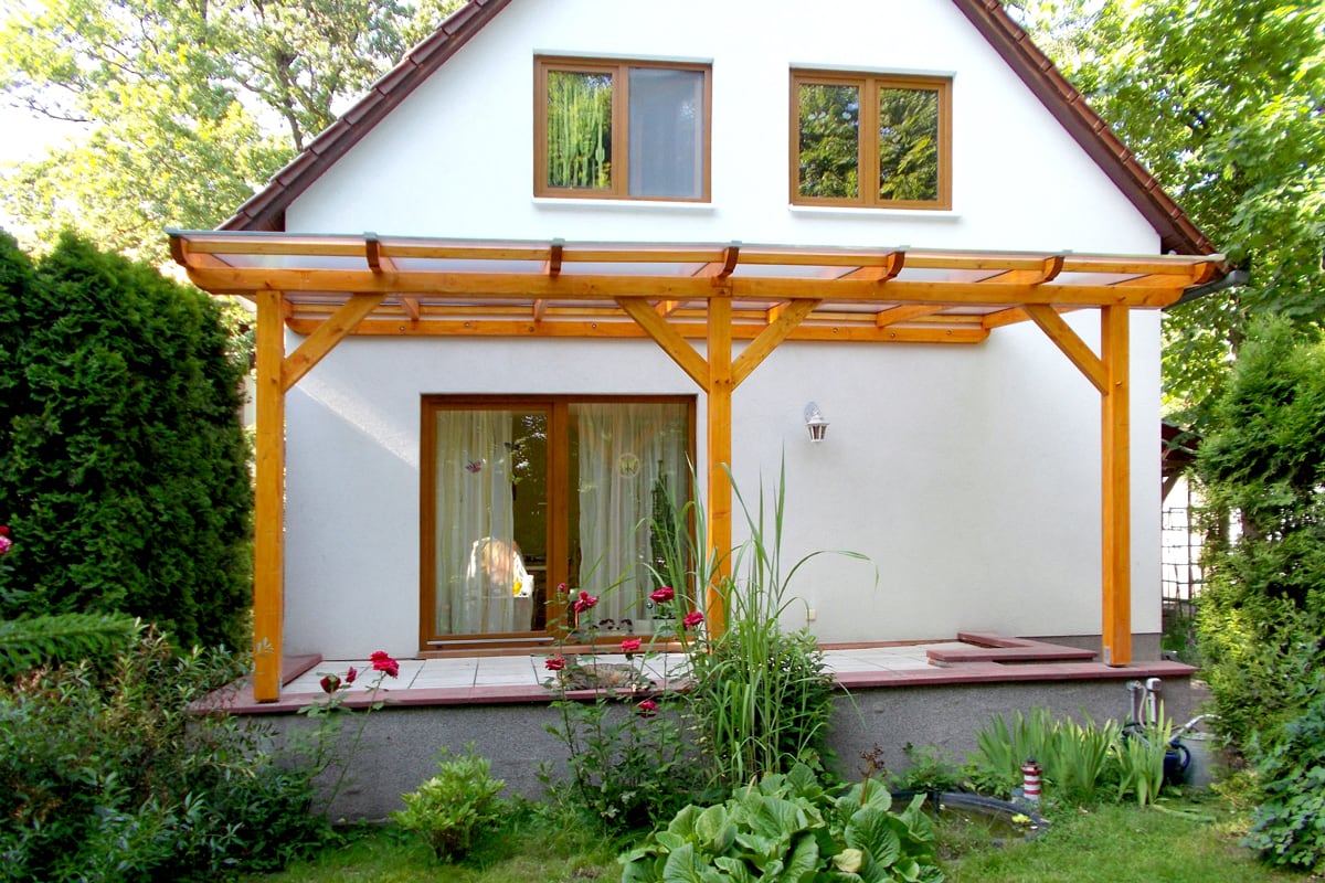 Eine schlichte Terrassenüberdachung aus hellem Holz erweitert das kleine Haus um einen geschützten Außenbereich. Umgeben von grünen Pflanzen und Rosen entsteht ein natürlicher und gemütlicher Rückzugsort.