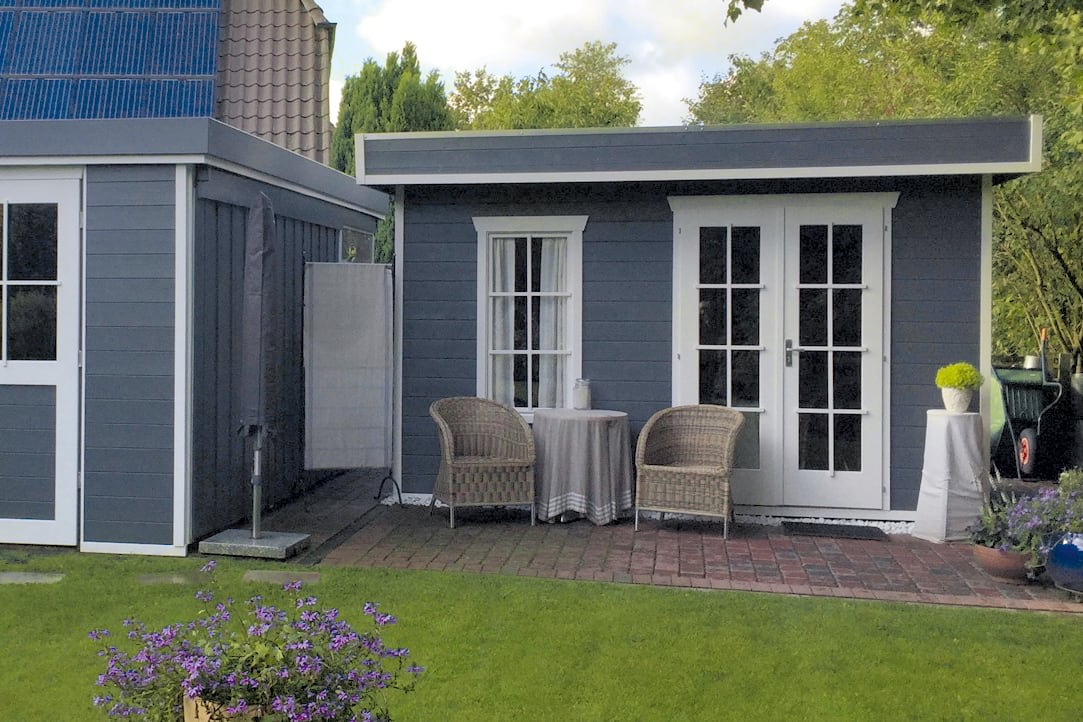Gartenhaus Breda mit zwei Korbsesseln und einem kleinen Tisch mit weißer Tischdecke auf gepflasterter Terrasse