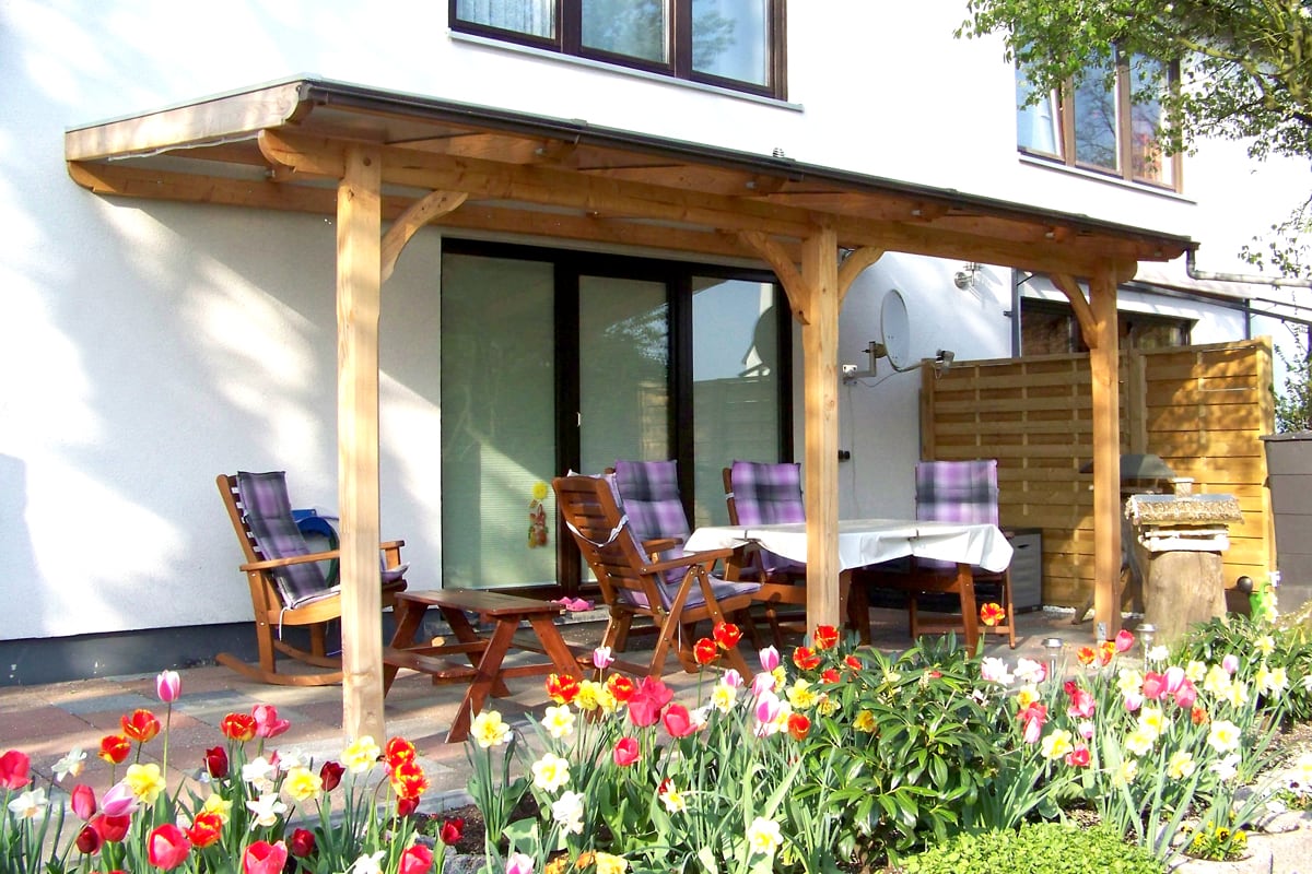 Eine überdachte Terrasse mit Holzkonstruktion, darunter Holzmöbel mit lila Sitzkissen und ein weiß gedeckter Tisch, im Vordergrund blühende bunte Tulpen.