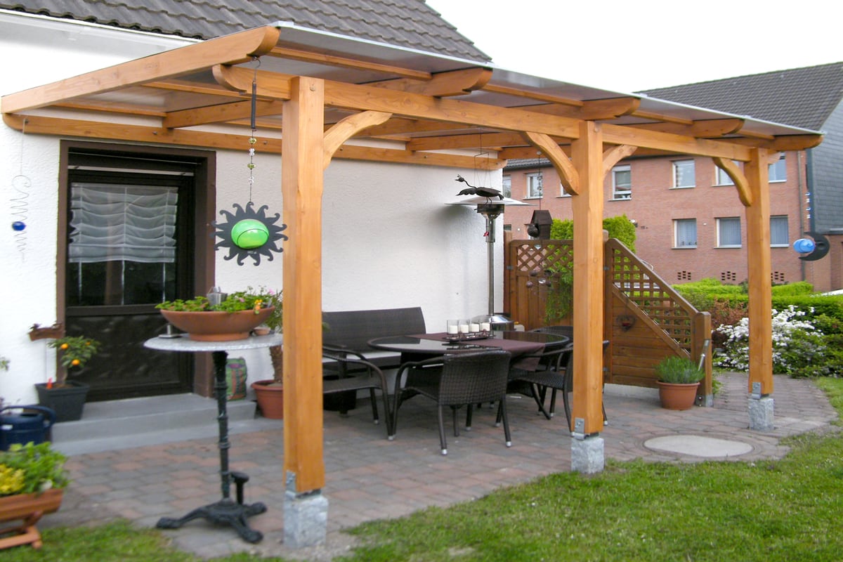 Eine überdachte Terrasse mit Holzkonstruktion, darunter ein Esstisch mit dunklen Rattan-Stühlen, dekoriert mit Pflanzen und Wanddeko, daneben ein kleiner Stehtisch mit Blumenschale.