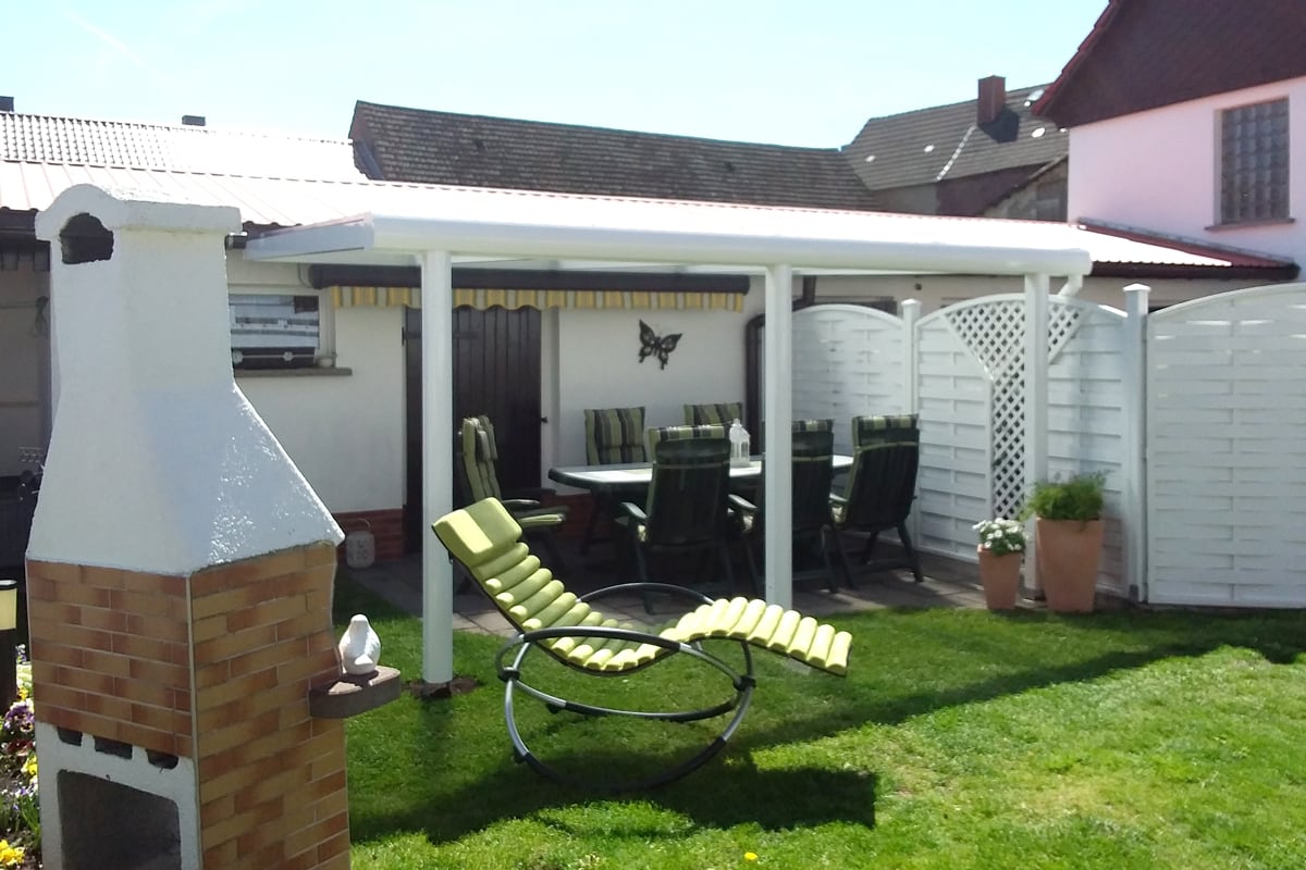 Weiße Terrassenüberdachung aus Aluminium, Tisch und Stühlen, davor gelb-grüner Liegestuhl auf Rasen, links gemauerter Grill.