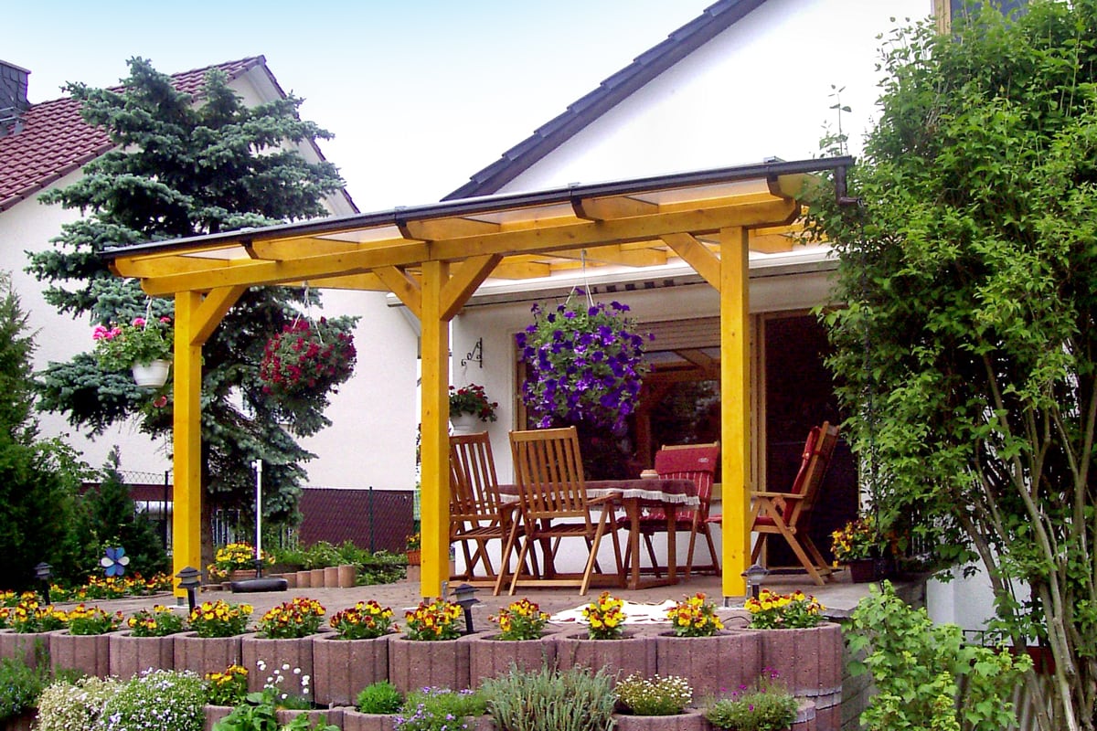 Eine eiche hell gestrichene Terrassenüberdachung aus Holz überdacht eine gemütliche Sitzgruppe auf der Terrasse mit Blick in den blühenden Garten.
