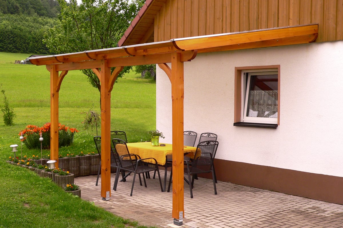 Eine überdachte Terrasse mit Holzkonstruktion, darunter ein Tisch mit gelber Tischdecke und schwarzen Metallstühlen, daneben ein Blumenbeet und grüne Wiese im Hintergrund.