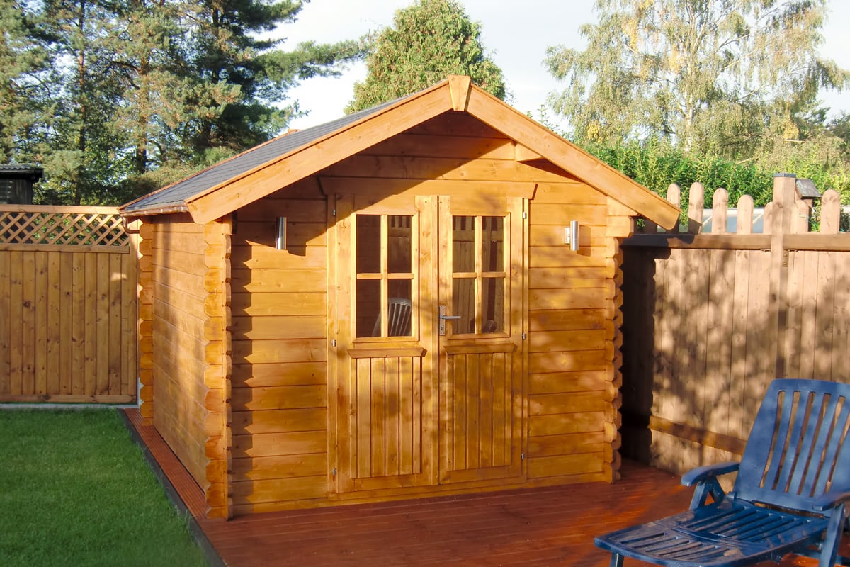 Holz-Gartenhaus Palma mit Doppeltür und Fenstern auf einer Holzterrasse, umgeben von Gartenzaun und Bäumen
