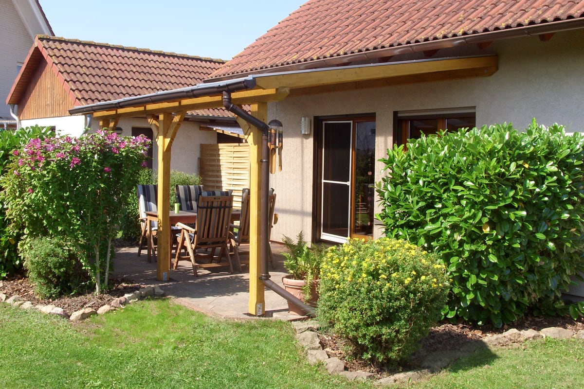 Eine kleine überdachte Terrasse mit Holzkonstruktion bietet einen gemütlichen Sitzplatz, umgeben von blühenden Sträuchern und viel Grün.