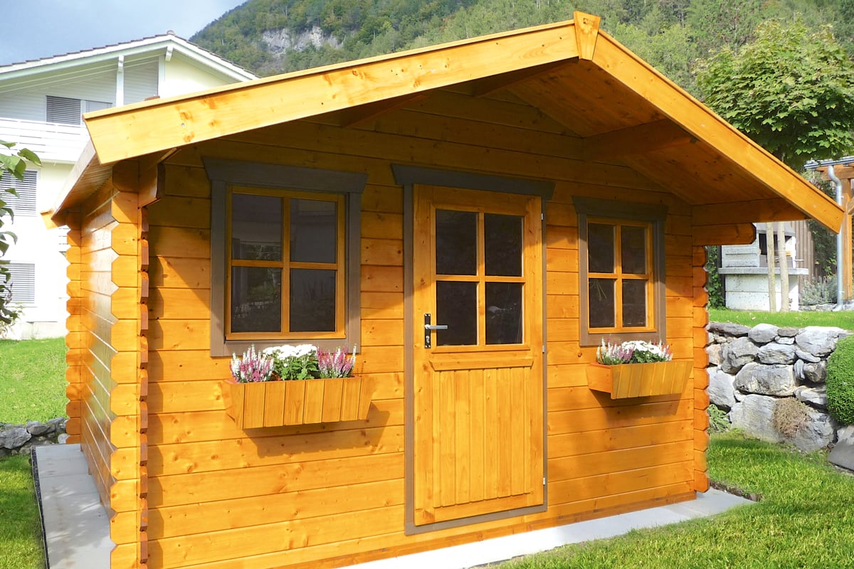 Holzhaus mit zwei Fenstern und einer Tür, Blumenkästen unter den Fenstern, im Garten vor Bergkulisse
