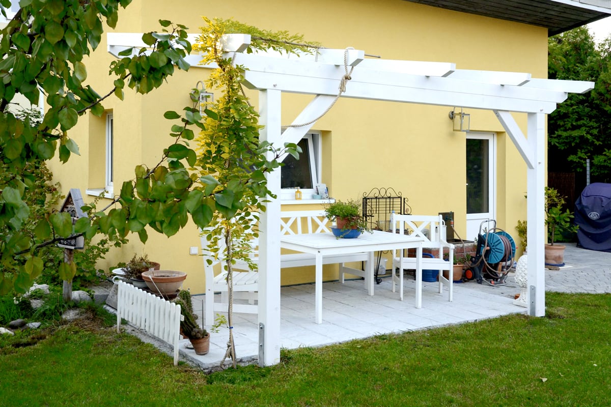 Eine weiße Pergola mit Sitzgarnitur schafft einen freundlichen Terrassenplatz vor dem gelben Haus.