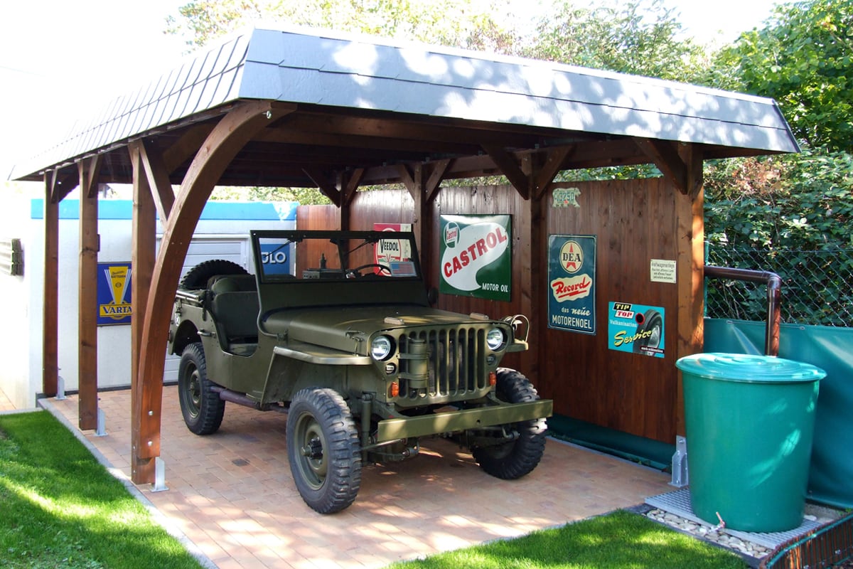 Offener Carport aus Holz mit Satteldach, darunter ein grüner Militärjeep auf gepflastertem Boden, umgeben von Rasen und Werbeschildern an der Rückwand.