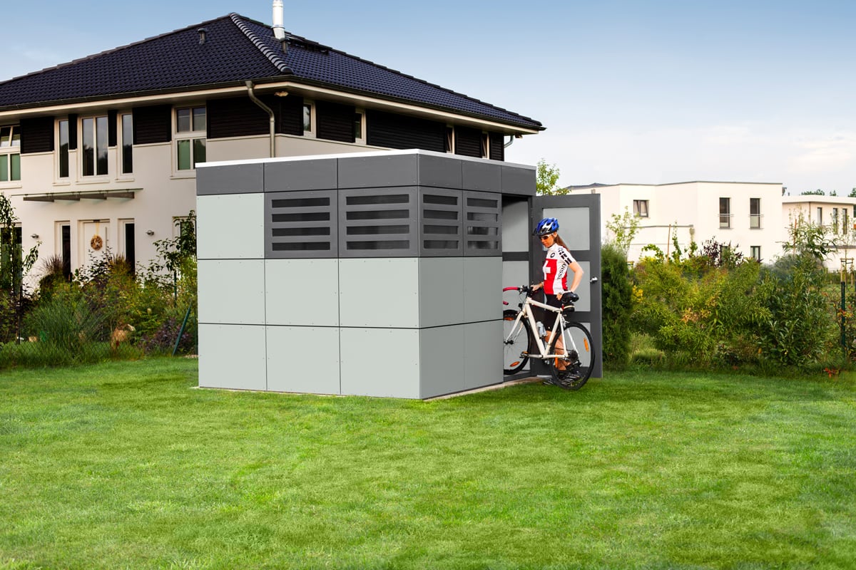 Modernes graues Gerätehaus CrossCube in Würfelform auf Rasen mit Sportlerin und Fahrrad