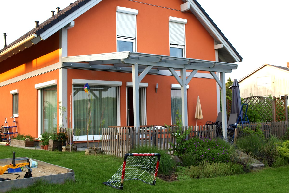 Eine helle Terrassenüberdachung aus Holz erweitert das orangefarbene Haus um einen geschützten Sitzplatz im Garten.