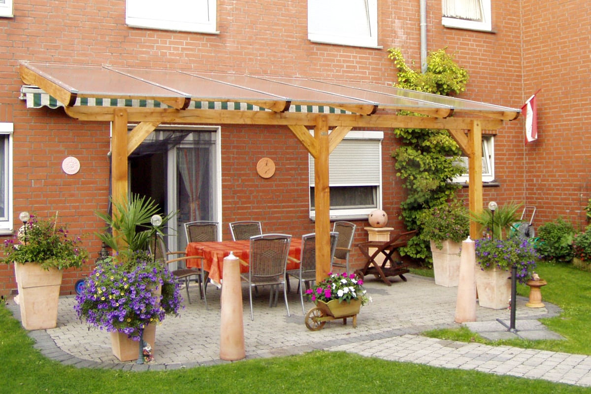 Eine Terrasse an einem roten Backsteinhaus mit Holzüberdachung, darunter ein Tisch mit orangefarbener Tischdecke und Stühlen, umrahmt von großen Blumenkübeln.