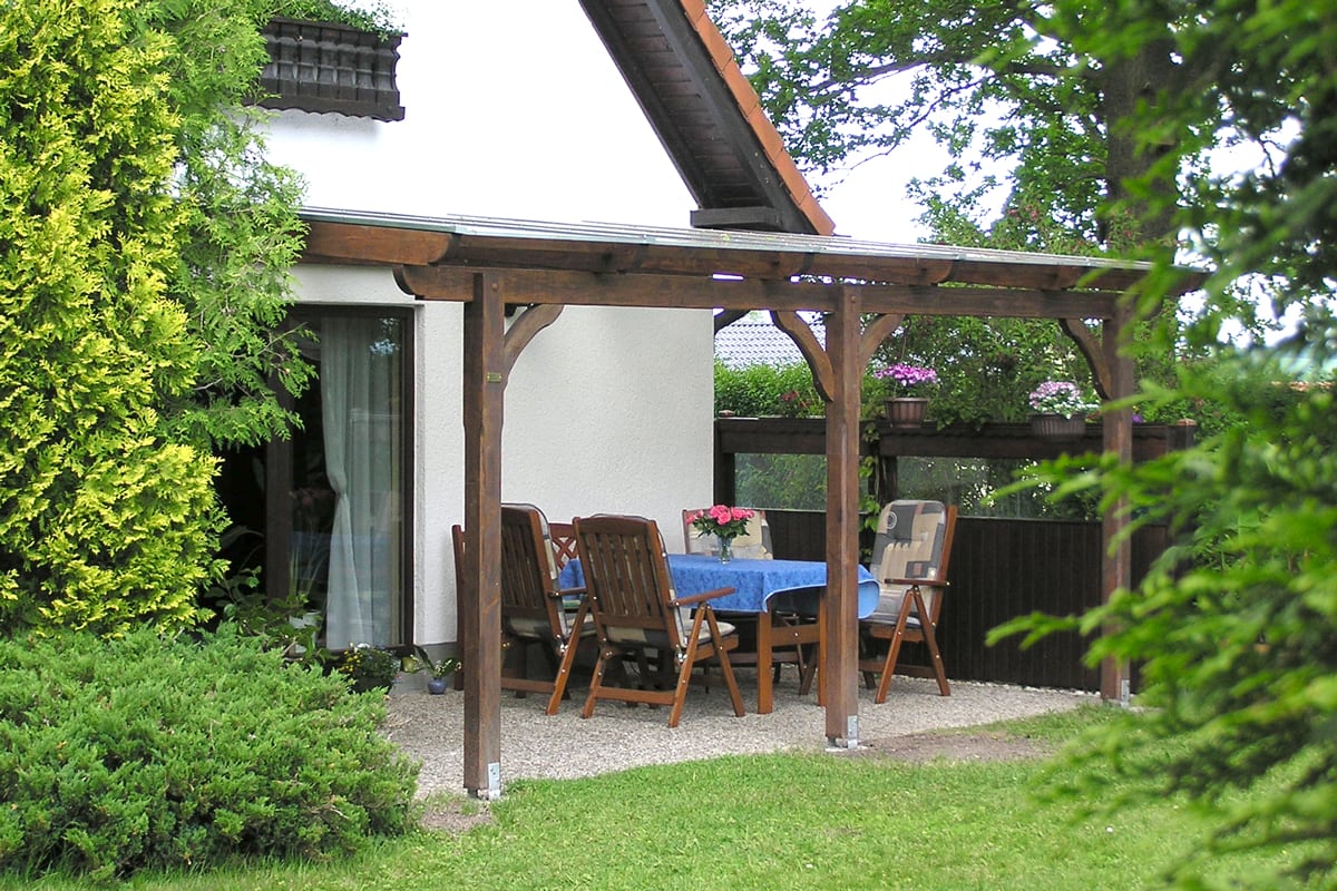 Eine Terrasse mit dunkler Holzüberdachung an einem weißen Haus, darunter ein Holztisch mit blauer Tischdecke und mehreren Stühlen, umgeben von Blumen und grünen Sträuchern.