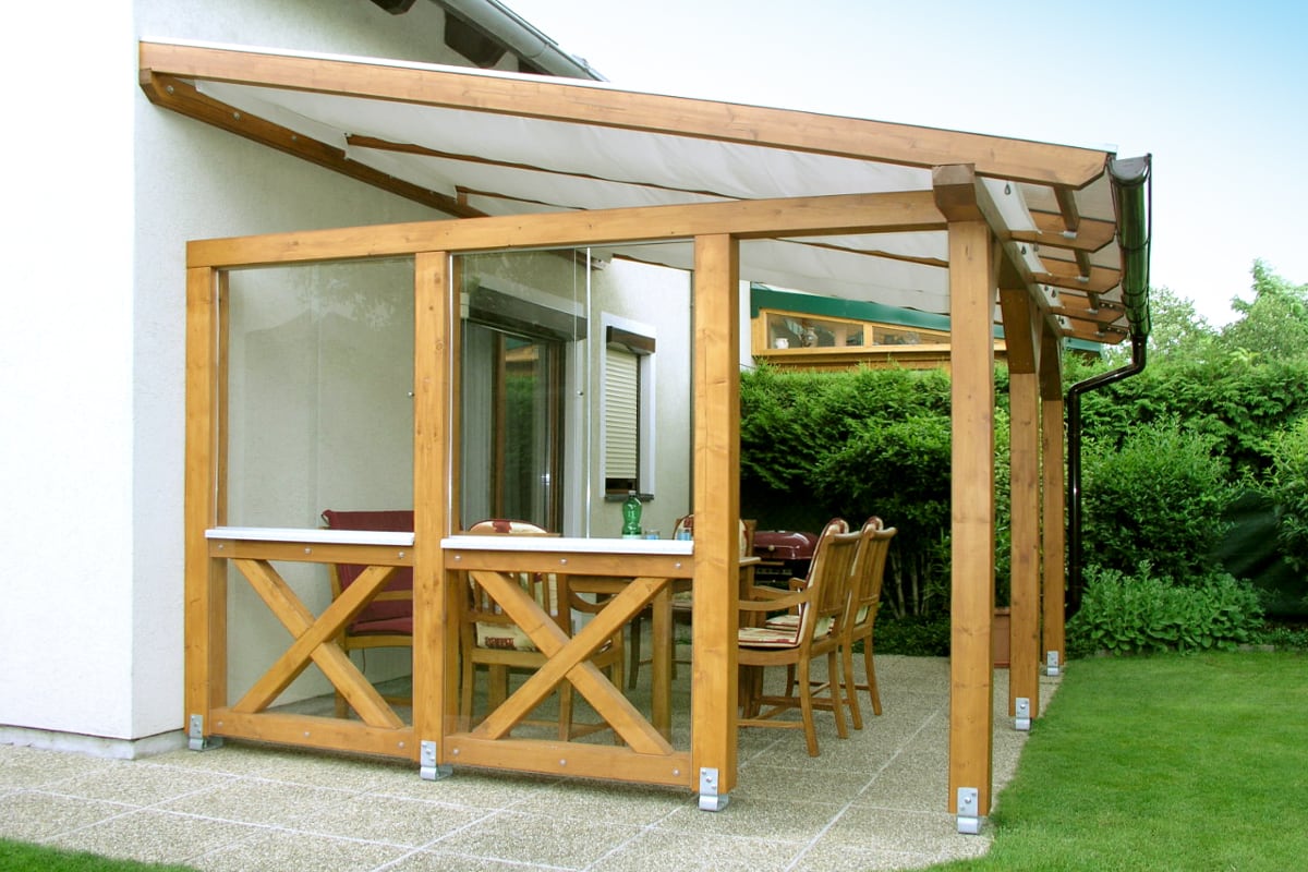 Eine Terrassenüberdachung aus Holz mit seitlichen Glaselementen überdacht eine geschützte Sitzecke direkt am Haus.