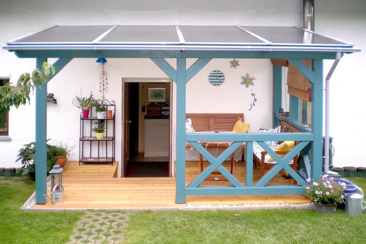 Eine türkis gestrichene Terrassenüberdachung aus Holz mit passendem Geländer überdacht eine gemütlich eingerichtete Sitzecke direkt am Haus.