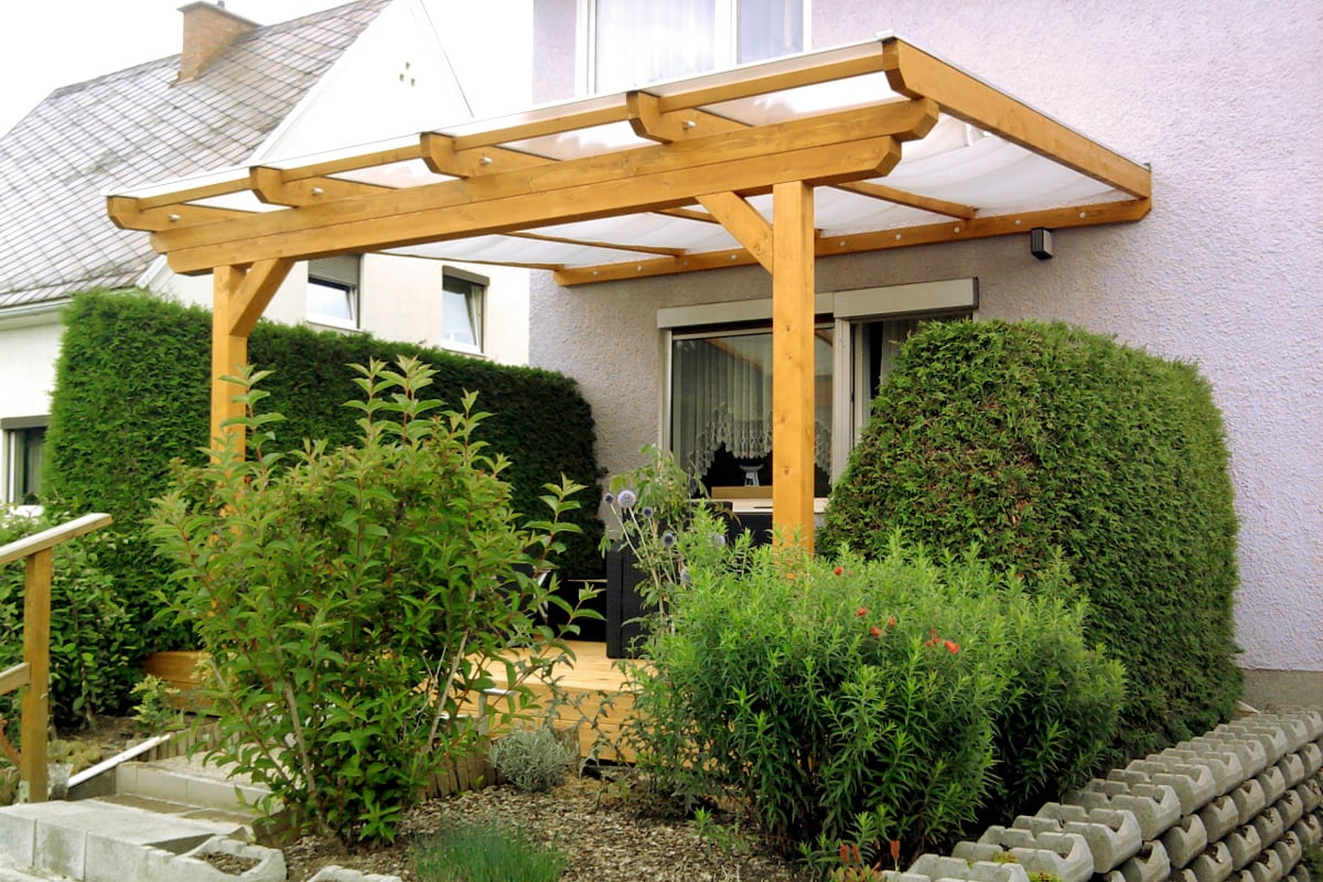 Ein massives Terrassendach aus naturbelassenem Holz fügt sich stabil an die Hauswand an. Die Konstruktion bietet mit ihrem klaren Design einen hellen und geschützten Platz im Grünen.