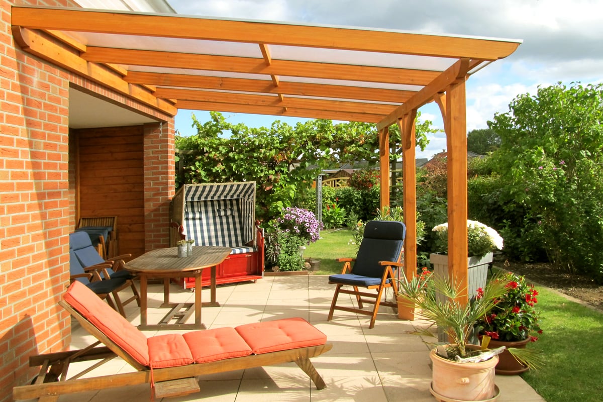 Eine helle Terrasse mit Holzüberdachung, darunter Gartenmöbel: ein Liegestuhl mit orangefarbenem Polster, ein blauer Stuhl, ein Holztisch und ein Strandkorb. Umgeben von Blumen und Pflanzen mit Blick in den grünen Garten.