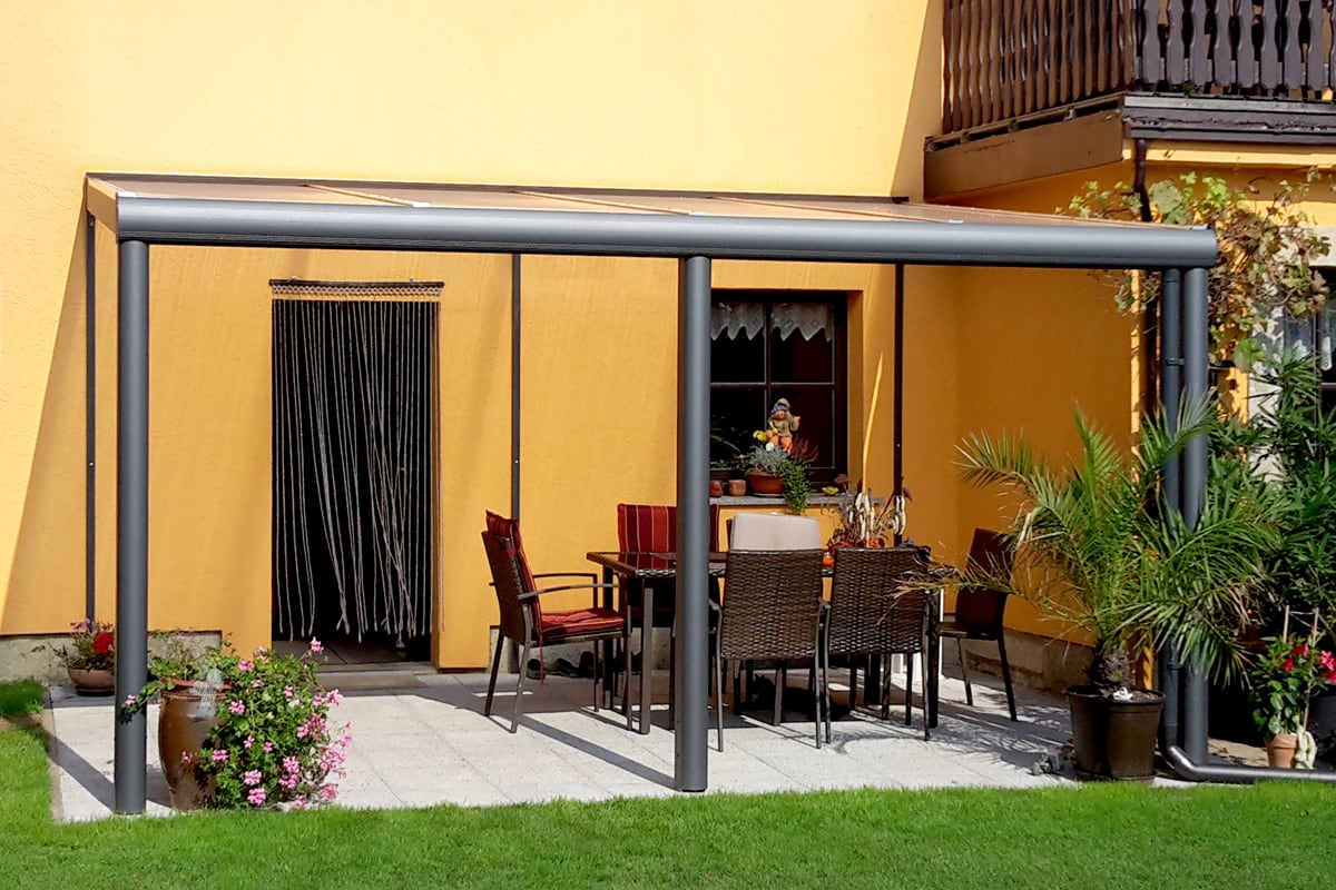 Terrassenüberdachung aus Aluminium in anthrazit, Tisch mit Stühlen, Pflanzen in Töpfen und gelber Hauswand im Hintergrund.