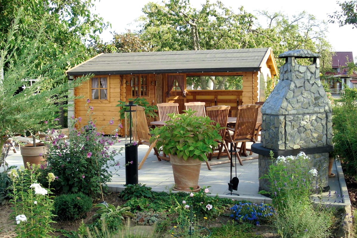 Garten mit Holzhaus Alicante , Terrasse mit Holztisch und Stühlen, großer Steingrill und blühenden Pflanzen