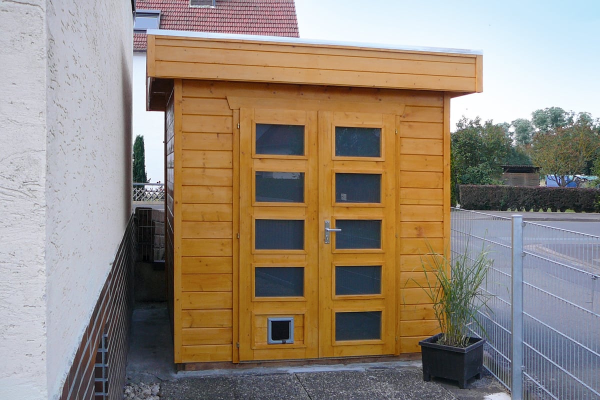 Kleines Gartenhaus Venlo aus Holz mit Doppeltür, vier Fenstern und Katzenklappe, neben Hauswand und Zaun