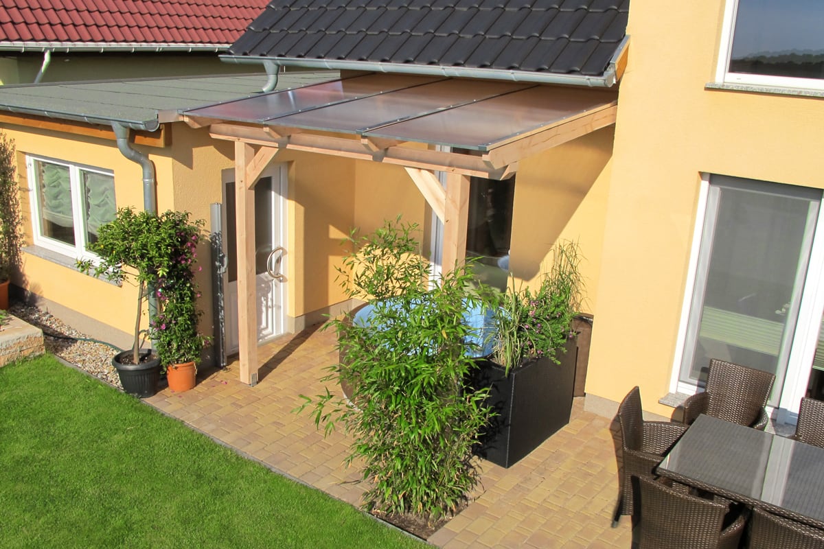Eine kleine überdachte Terrasse mit heller Holzkonstruktion bietet neben dem Haus einen geschützten Bereich mit Pflanzen und Sitzmöbeln.