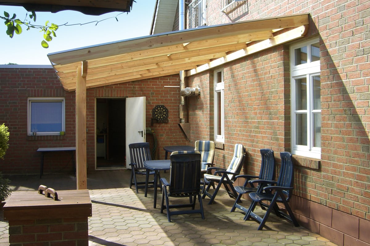 Eine helle Holzüberdachung an einer Backsteinwand, darunter mehrere dunkelblaue Gartenstühle mit Polstern und ein kleiner Tisch auf gepflastertem Boden.