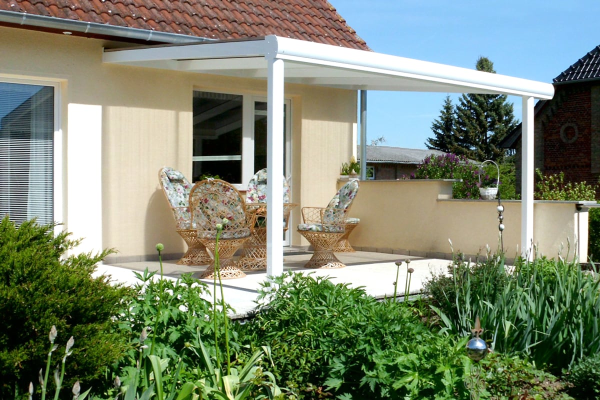 Weiße Terrassenüberdachung aus Aluminium, zwei geflochtenen Stühlen mit floral gemusterten Polstern vor einem hellen Haus, umgeben von grünem Garten.