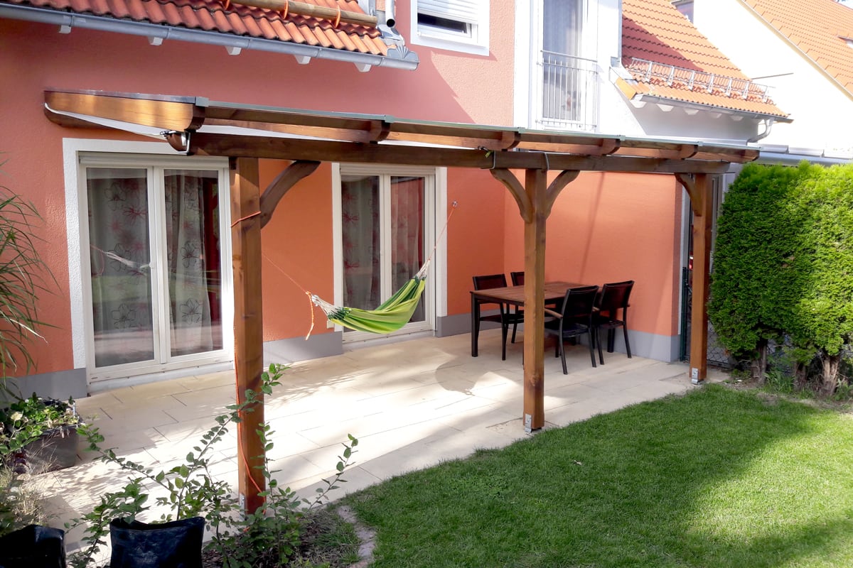Eine überdachte Terrasse mit Holzkonstruktion an einem Haus mit orangefarbener Fassade. Unter der Überdachung hängt eine grün-gelbe Hängematte, daneben steht ein Tisch mit schwarzen Stühlen. Im Vordergrund ist eine Rasenfläche und seitlich Hecken.