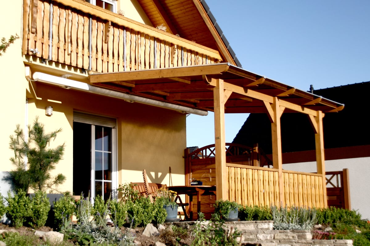 Eine erhöhte Holzterrasse mit Überdachung und seitlicher Holzbrüstung an einem gelben Haus mit Balkon, umgeben von bepflanzten Beeten.