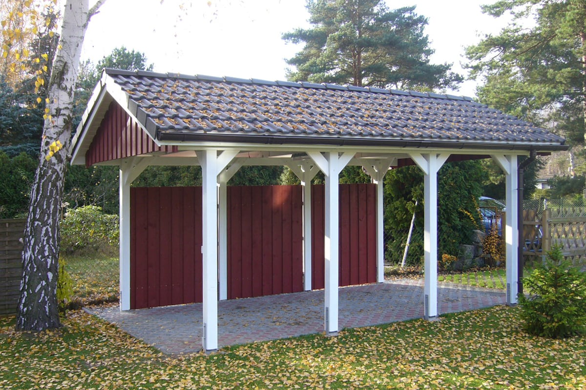 Freistehende, offene Holzgarage mit weiß gestrichenen Stützpfeilern und rotbraunen Holzpaneelen, mit Ziegeldach, umgeben von herbstlichem Laub und Bäumen.