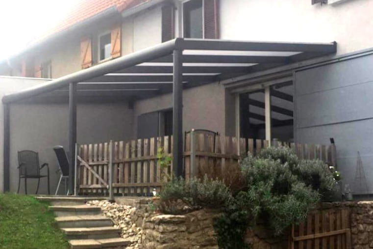 Terrassenüberdachung aus Aluminium in anthrazit, Holzgeländer und Stufen aus Stein, umgeben von Sträuchern und Rasen.
