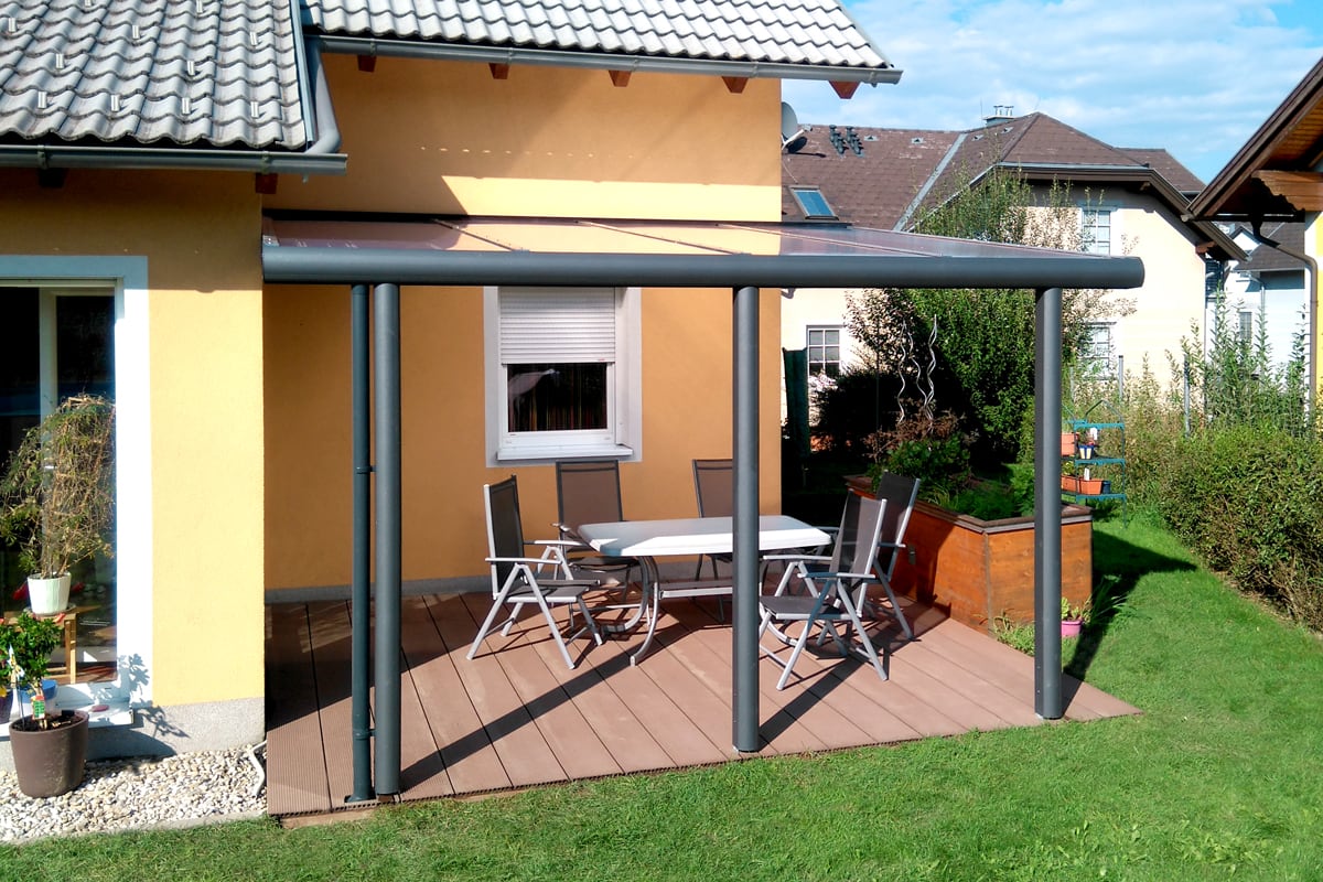 Moderne Terrassenüberdachung mit pulverbeschichtetem Aluminiumgestell und klarer Dacheindeckung für optimalen Wetterschutz. Die schlanke Konstruktion fügt sich harmonisch in die Hausarchitektur ein und bietet einen geschützten Platz im Freien.