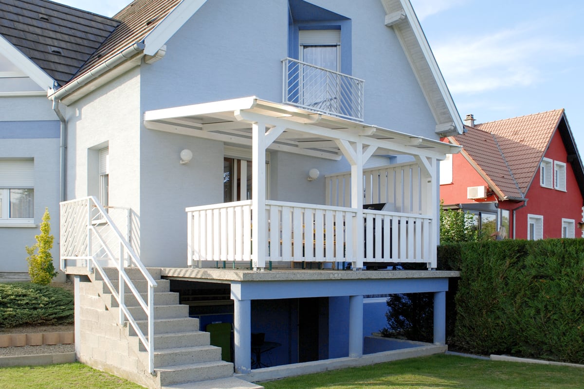Eine weiß gestrichene Terrassenüberdachung aus Holz mit passendem Geländer überdacht den erhöhten Balkon eines Einfamilienhauses.