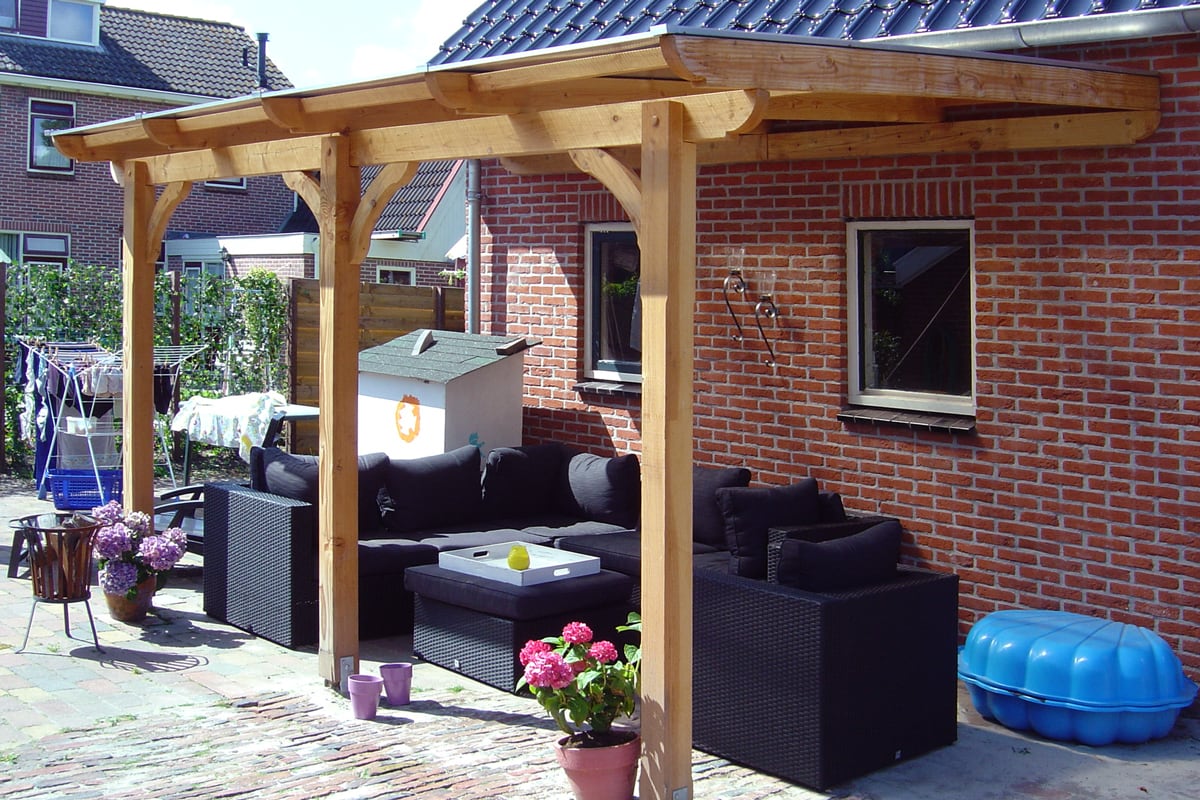 Eine überdachte Terrasse mit Holzkonstruktion, schwarzer Rattan-Sofalandschaft und Pflanzkübeln vor einer roten Backsteinwand.