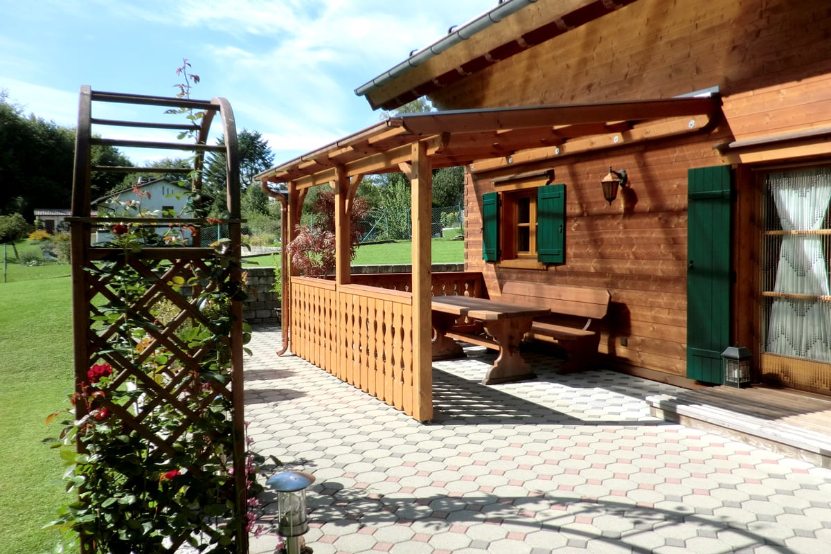 Eine Holzterrasse mit Überdachung an einem Blockhaus mit grünen Fensterläden; darunter steht eine rustikale Sitzbank mit Tisch, davor eine gepflasterte Fläche mit Rankgitter und blühenden Pflanzen.