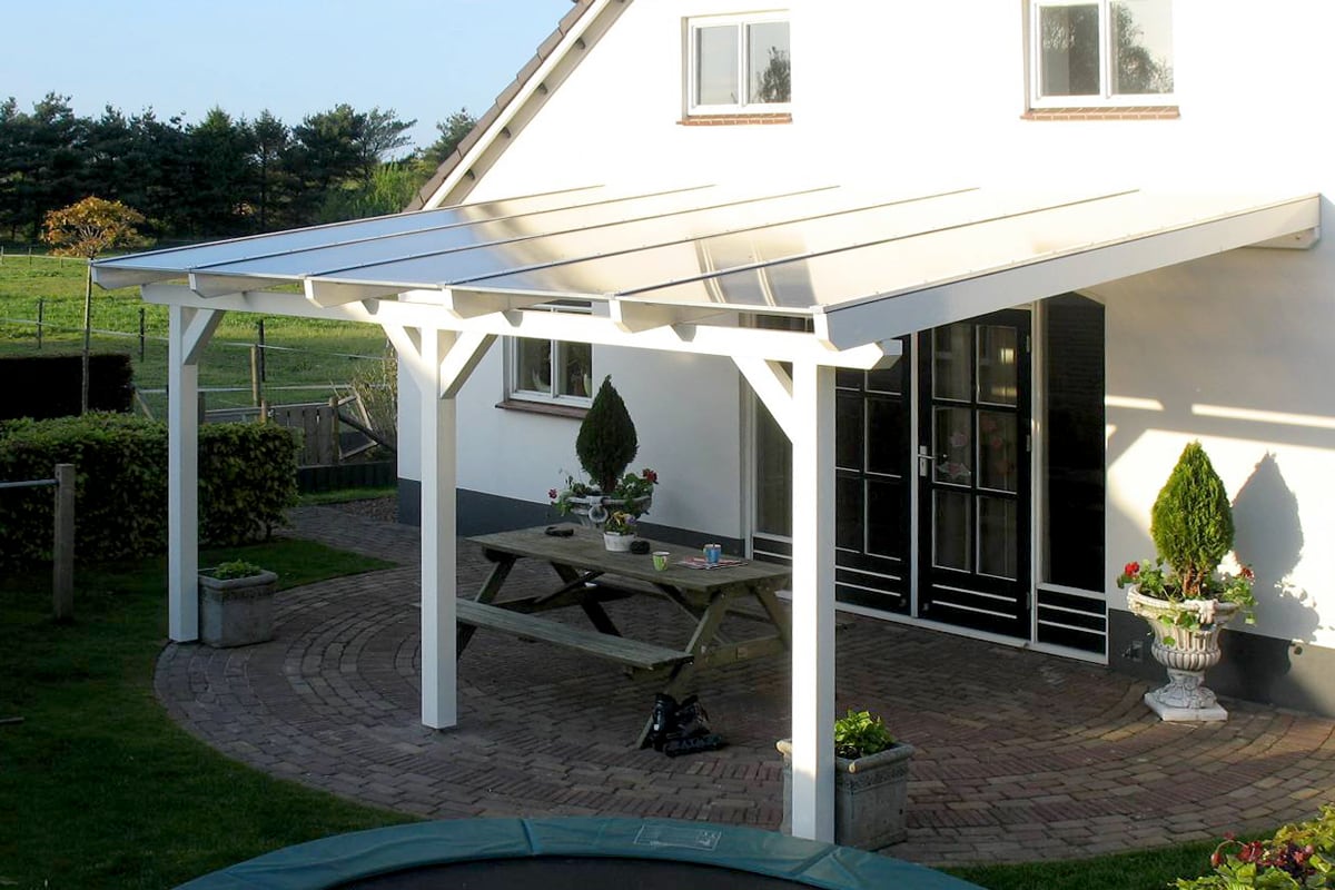 Eine weiß gestrichene Terrassenüberdachung aus Holz mit Glasdach schützt eine Terrasse mit Picknicktisch und Blick ins Grüne.