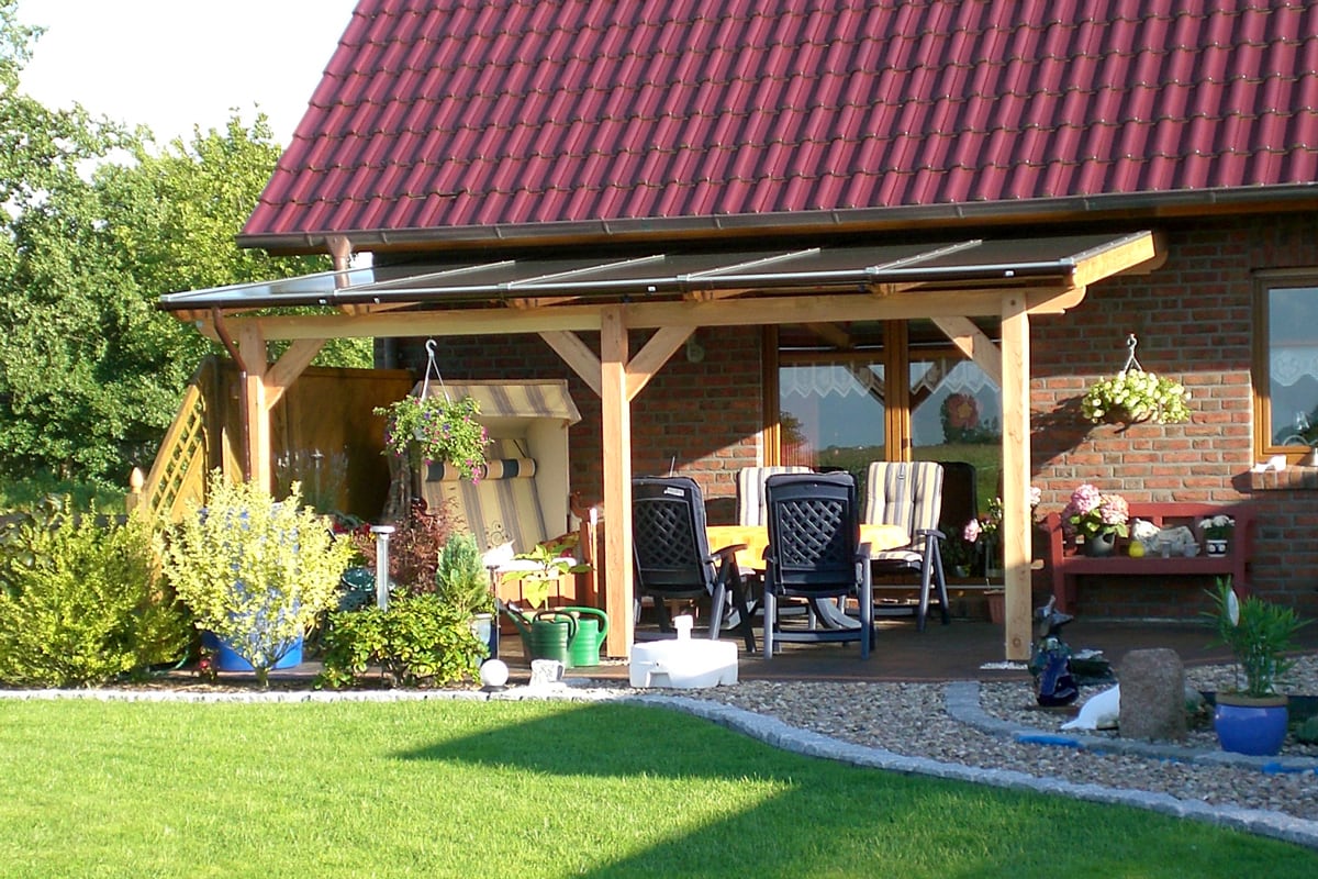 Ein Haus mit rotem Ziegeldach und Backsteinfassade, davor eine Holzterrassenüberdachung mit transparentem Dach. Darunter stehen Gartenmöbel mit Polstern und ein Strandkorb, umgeben von vielen Pflanzen und Blumen.
