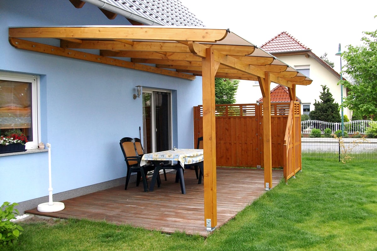 Eine überdachte Terrasse mit Holzgestell und durchsichtigem Dach an einem hellblauen Haus. Darunter stehen ein Tisch mit Wachstuchdecke und Stühle, der Boden ist aus Holz verlegt, und ein Sichtschutz aus Holz grenzt den Bereich zur Straße ab.