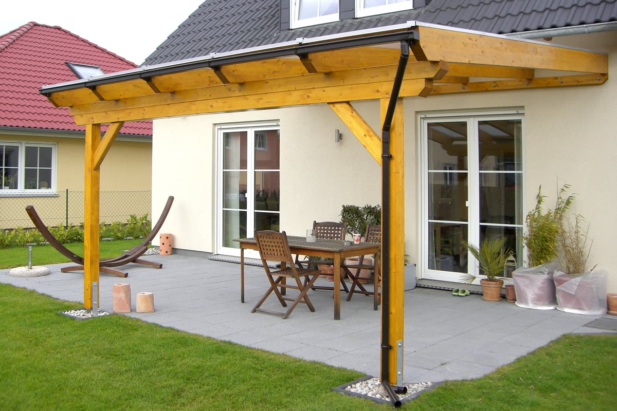 Eine moderne Terrassenüberdachung aus Holz bietet Schutz direkt am Haus und schafft einen gemütlichen Essplatz im Freien. Schlichte Gartenmöbel und einige Pflanzen runden das einladende Ambiente ab.