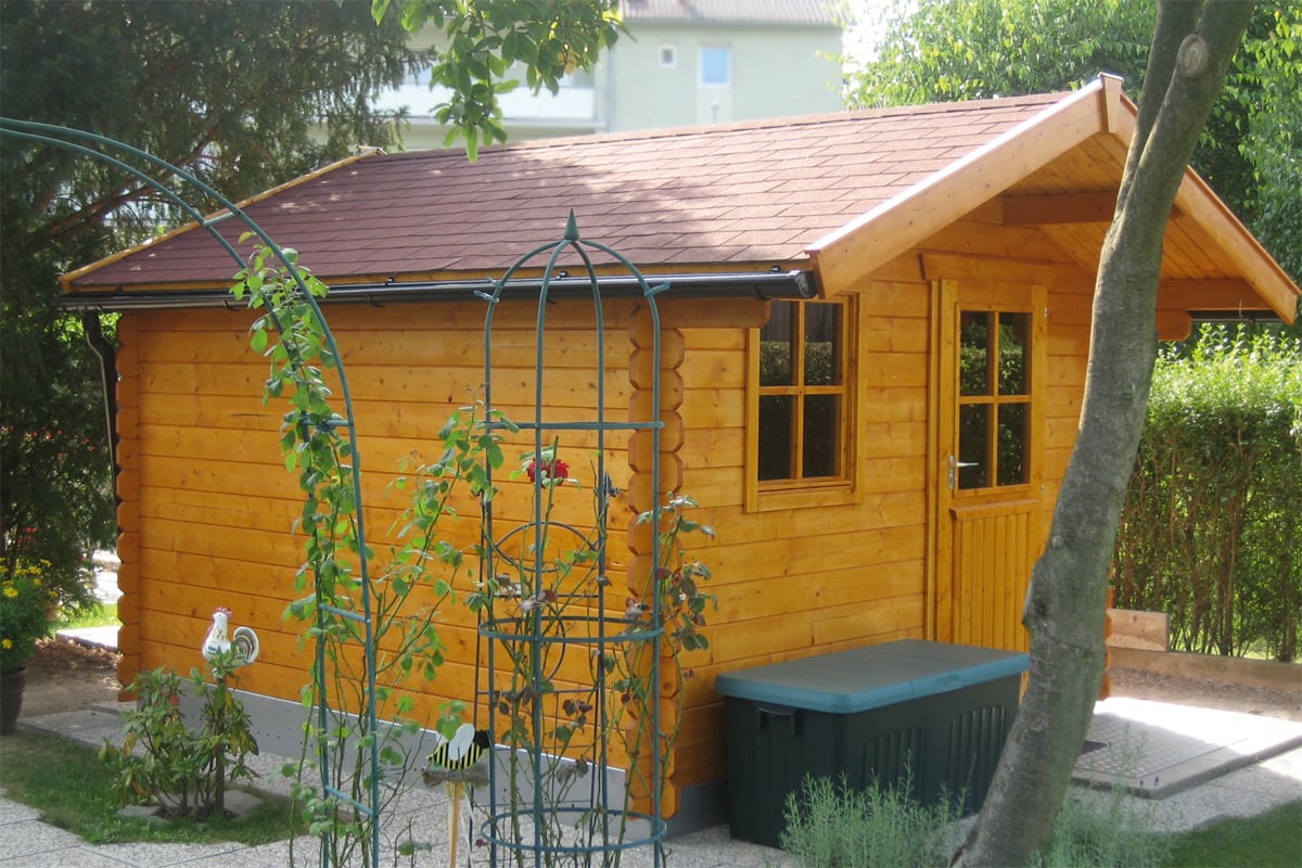 Holz-Gartenhaus mit zwei Fenstern und einer Tür, umgeben von Pflanzen und einem Baum