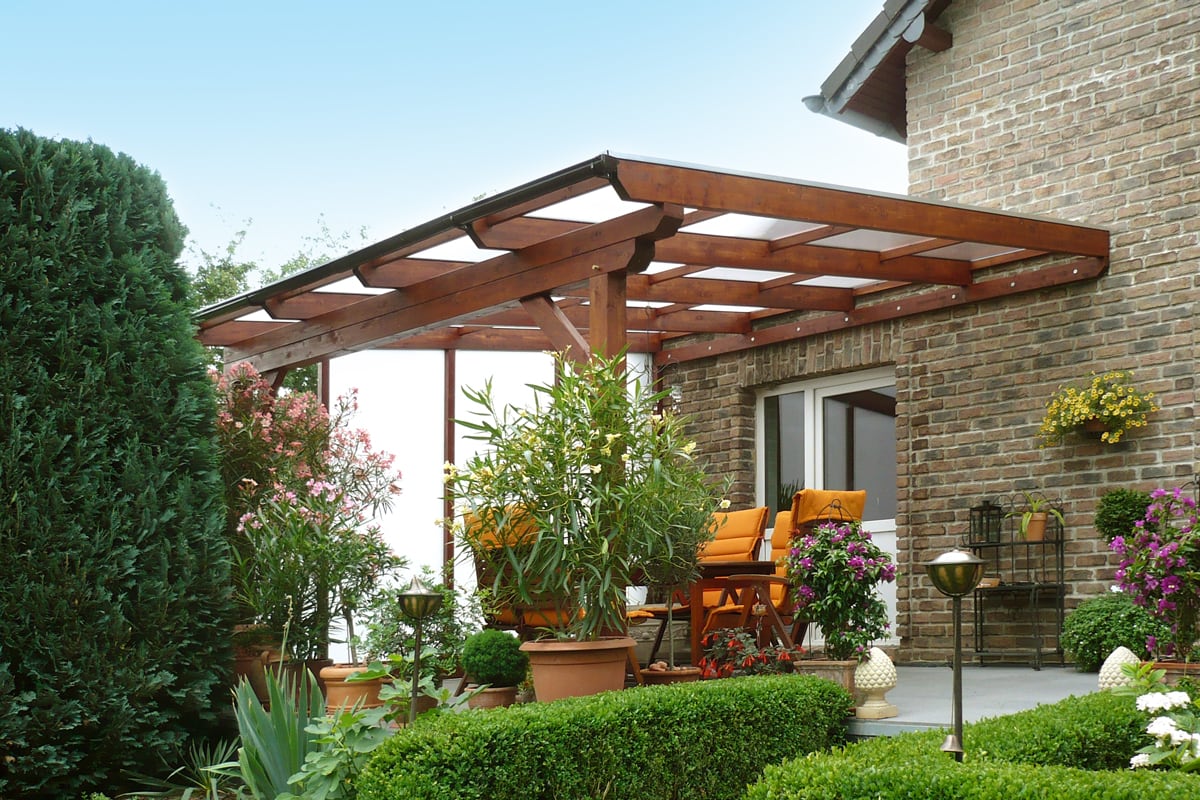 Eine überdachte Terrasse mit Holzkonstruktion und vielen blühenden Pflanzen bietet einen farbenfrohen und geschützten Sitzbereich am Haus.