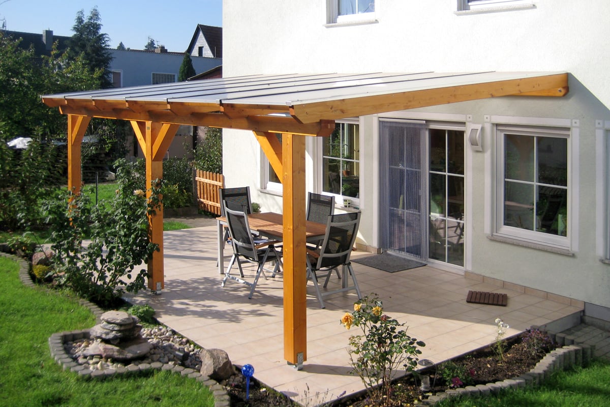 Eine helle Terrassenüberdachung aus Holz schützt den gefliesten Sitzbereich mit Tisch und Stühlen im Garten.