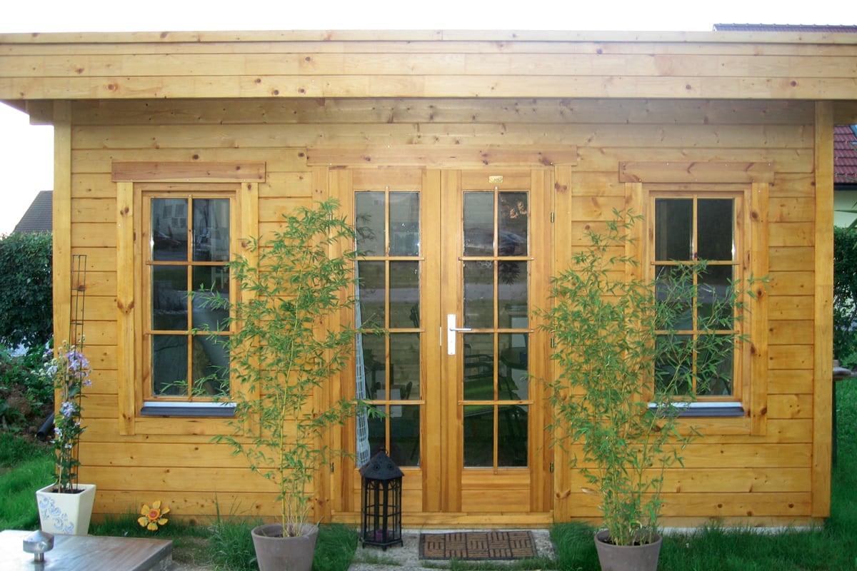 Holzhaus Breda mit zwei Fenstern und einer Glastür, davor drei Pflanzen in Töpfen und eine Laterne