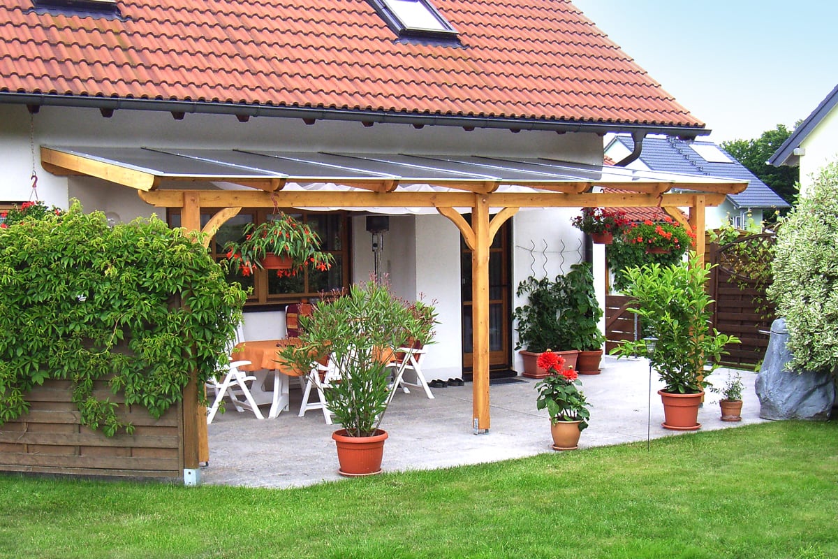 Eine überdachte Terrasse mit Holzkonstruktion an einem Haus mit rotem Ziegeldach; darunter stehen weiße Gartenmöbel mit orangener Tischdecke, umgeben von vielen Topfpflanzen und hängenden Blumentöpfen.