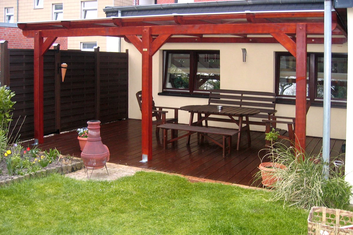 Eine überdachte Terrasse mit dunkelroter Holzkonstruktion und Holzboden, darauf eine Sitzgruppe aus Holzbänken und Tisch, daneben Pflanzenkübel und ein Terrassenofen.