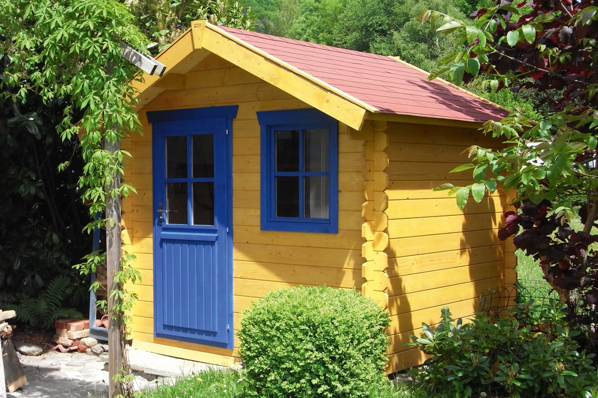 Gelbes Gartenhaus mit blauer Tür und Fenstern, rotem Satteldach, umgeben von grünen Pflanzen