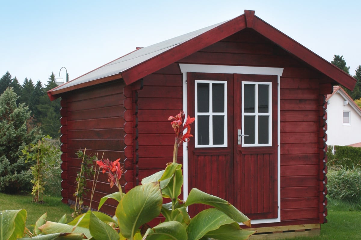 Kleines rotes Gartenhaus Palma mit weißen Fensterrahmen und Doppeltür, umgeben von grünen Pflanzen