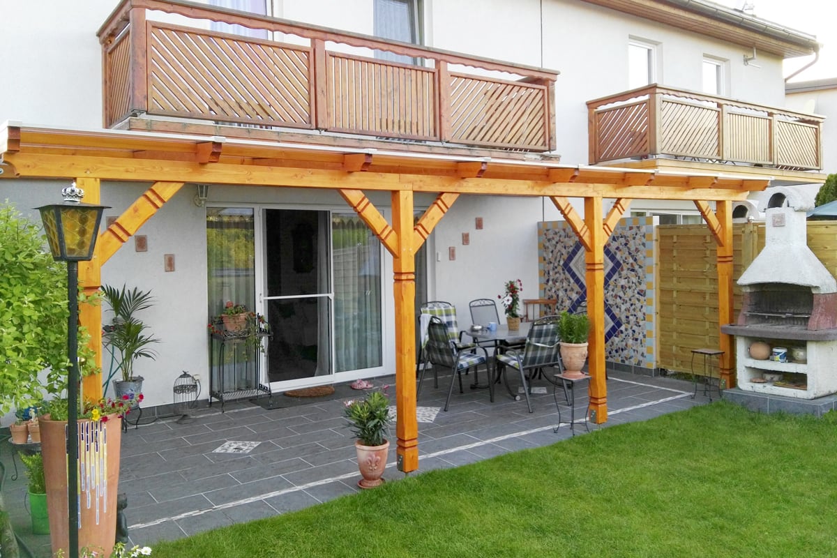 Eine helle Holzüberdachung schützt die Terrasse mit Sitzbereich und festem Grillplatz im Garten.