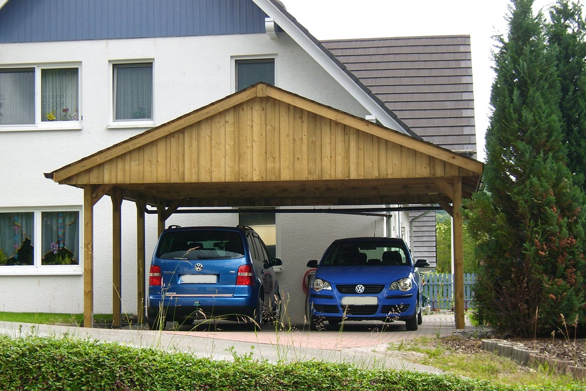 Holzcarport mit Satteldach, das zwei blaue Volkswagen-Pkw schützt, vor einem weißen Wohnhaus mit grauem Dach.